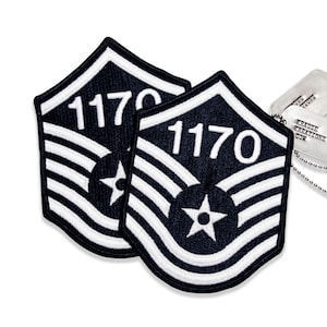May include: Two black and white embroidered patches with the number "1170" and a star emblem. The patches are in the shape of a chevron. A silver dog tag with the text "KRAUSE KREATIONS COR" is visible in the background.