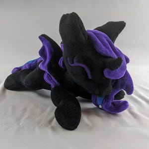 Puede incluir: Un dragón de peluche con pelaje negro y morado, acostado de lado con la cabeza apoyada en su pata. El dragón tiene una gema azul en su pecho.