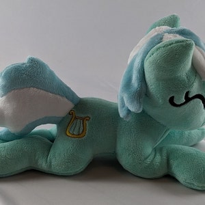 Pu&ograve; includere: Un giocattolo di peluche di un pony blu chiaro e bianco con una lira dorata su un lato. Il pony &egrave; sdraiato su un fianco con la testa appoggiata sulla zampa anteriore.