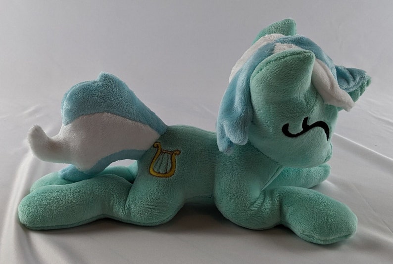 Pu&ograve; includere: Un giocattolo di peluche di un pony blu chiaro e bianco con una lira dorata su un lato. Il pony &egrave; sdraiato su un fianco con la testa appoggiata sulla zampa anteriore.
