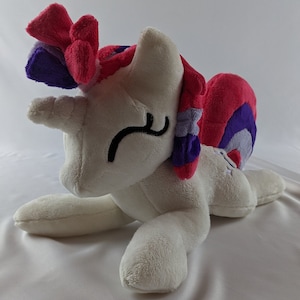 May include: A white plush unicorn with a pink, purple and white mane and tail. The unicorn is lying on its side with its head resting on its front leg. It has a closed eye and a small smile.