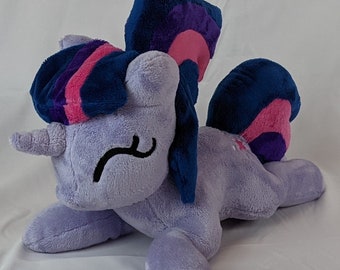 May include: A plush toy of Twilight Sparkle from My Little Pony. The toy is purple with a blue mane and tail. The mane is styled in a high ponytail with a pink streak. The toy is lying on its side with its head resting on its front paw.