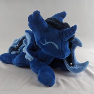 Puede incluir: Un juguete de peluche azul en forma de unicornio con una luna creciente en su flanco. El unicornio tiene una melena y una cola azul oscuro, y un cuerpo azul claro. El juguete está acostado de lado con la cabeza apoyada en su pata delantera.