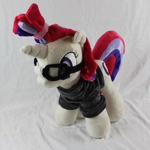 May include: A white plush toy unicorn with pink and purple hair, wearing black goggles and a black sweater. The unicorn has a pink and purple tail and a horn.