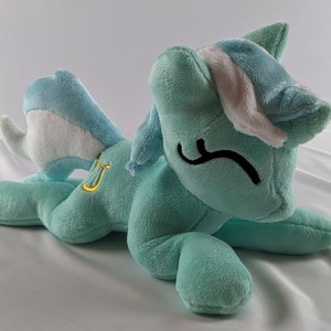 Pu&ograve; includere: Un giocattolo di peluche a forma di pony blu chiaro con criniera e coda bianche. Il pony indossa una fascia bianca con una striscia blu chiaro e un simbolo di lira dorato sul lato.