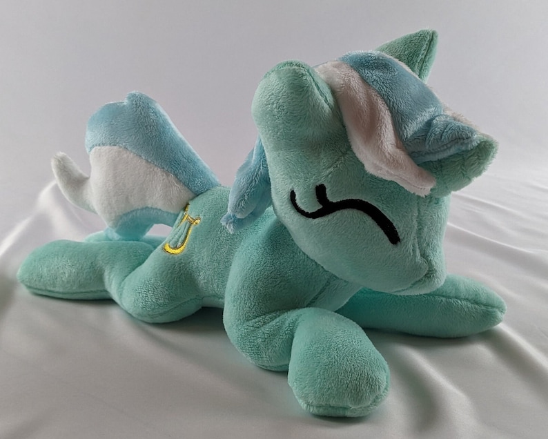 Pu&ograve; includere: Un giocattolo di peluche a forma di pony blu chiaro con criniera e coda bianche. Il pony indossa una fascia bianca con una striscia blu chiaro e un simbolo di lira dorato sul lato.