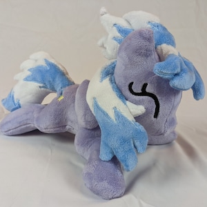 May include: A plush toy of a purple and blue pony with a white mane and tail. The pony is lying on its side with its head turned to the side. The pony has a black eye and a black swirl on its cheek.