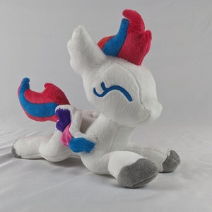 Puede incluir: Juguete de peluche blanco con una melena y cola rosa, azul y morada. El juguete tiene una sonrisa bordada azul y está acostado de lado.