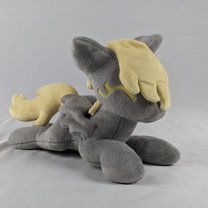 Puede incluir: Un juguete de peluche gris con una melena y una cola amarillas. El juguete está en posición sentada con la cabeza inclinada hacia un lado.