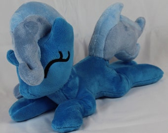 May include: A plush toy of a blue pony with a light blue mane and tail. The pony is lying on its side with its head resting on its front leg.