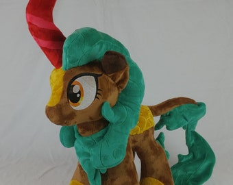 Può includere: Un giocattolo di peluche di un pony marrone con criniera e coda verdi. Il pony ha un corno rosso e segni gialli. Il pony è in piedi su uno sfondo bianco.