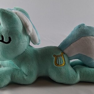 Pu&ograve; includere: Un pony di peluche blu chiaro con criniera e coda bianche. Il pony ha una lira dorata ricamata su un lato.