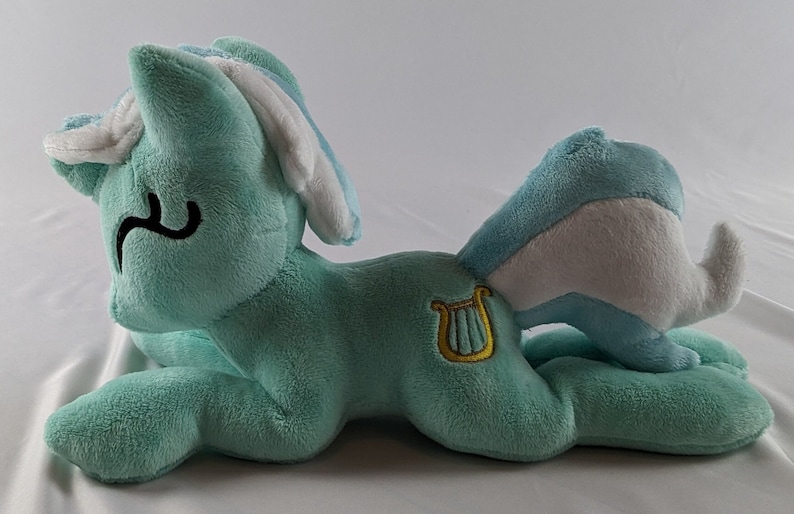 Pu&ograve; includere: Un pony di peluche blu chiaro con criniera e coda bianche. Il pony ha una lira dorata ricamata su un lato.