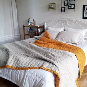 Puede incluir: Una cama blanca con una cabecera blanca y una colcha gris y amarilla. La cama está hecha con sábanas y almohadas blancas. Hay una mesita de noche blanca junto a la cama con un pequeño espejo en la parte superior.