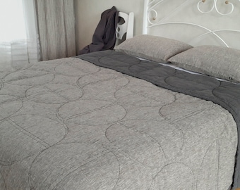 May include: A bed with a white metal headboard and a gray quilt with a circular stitched pattern. The quilt has a dark gray border. Two gray pillows rest on the bed, which is covered with a white sheet.