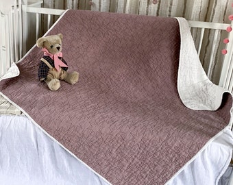 May include: A mauve quilted baby blanket with a white border, featuring a teddy bear sitting on top. The blanket is on a white crib with a white ruffle skirt. The crib is in a nursery with a white dresser and a white stool.