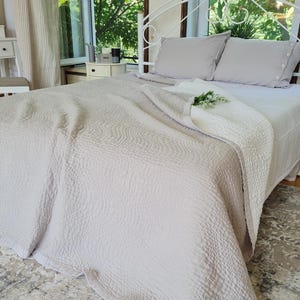 Puede incluir: Un cubrecama de tonos neutros con una superficie texturizada cubre una cama con almohadas grises. La cama está colocada contra un marco de metal blanco. Una alfombra a juego está en el suelo. La habitación tiene un ambiente luminoso y aireado.