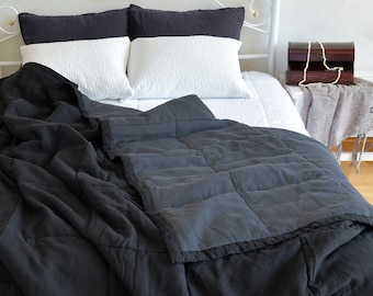 May include: A black quilted linen throw blanket with a white bed in the background. The blanket is folded and draped over the bed.
