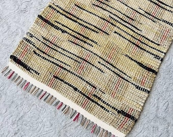 May include: A rectangular woven runner rug featuring a textured pattern of tan, beige, and cream fibers. The design includes horizontal streaks of black and thin red accents throughout the weave, finished with short, multicolored fringe at both ends.