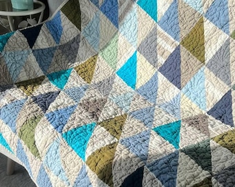 May include: A handmade quilt with a geometric pattern of triangles in shades of blue, green, and gray. The quilt is draped over a gray sofa, showcasing its textured surface and detailed stitching. The quilt's design adds a touch of modern style.