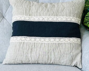 May include: A square throw pillow featuring a quilted beige fabric base with a wide horizontal black band across the center. The black section is bordered by two strips of intricate white lace trim, creating a distinct contrast against the neutral quilted background.