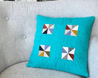 May include: A turquoise square pillow with a quilted pattern and four square patchwork designs. Each patchwork square features a pinwheel design in various colors, including red, black, white, and yellow. The pillow is on a gray armchair.