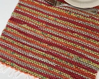 May include: A rectangular woven placemat featuring a multi-colored striped pattern in shades of red, orange, yellow, and green. The textile has a textured, nubby surface and is finished with short white fringe along the bottom edge.