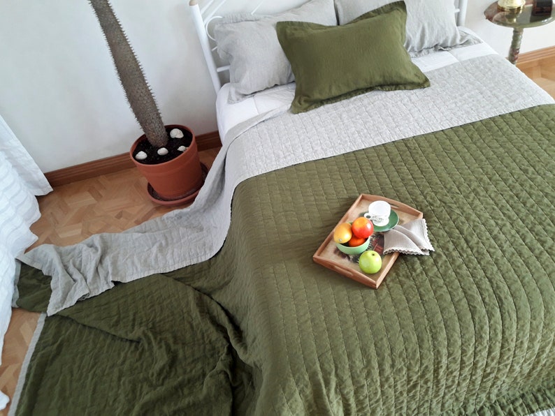 Puede incluir: Una cama con ropa de cama verde oliva y gris. La cama tiene una colcha verde oliva acolchada y una almohada a juego. Una bandeja de madera con fruta y una taza de t&eacute; est&aacute; sobre la cama. Un cactus en maceta est&aacute; al lado de la cama.