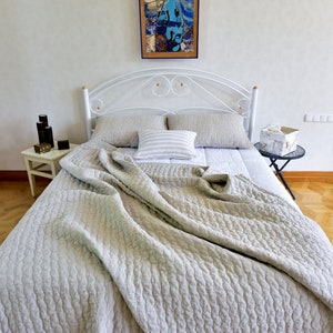 K&ouml;nnte beinhalten: Ein wei&szlig;es Metallbettgestell mit einer hellgrauen Leinenbettdecke und zwei Kissen. Das Bett ist mit einem wei&szlig;en Laken und einem gestreiften Kissen bezogen. Das Bett steht in einem Zimmer mit wei&szlig;en W&auml;nden und einem Holzboden.