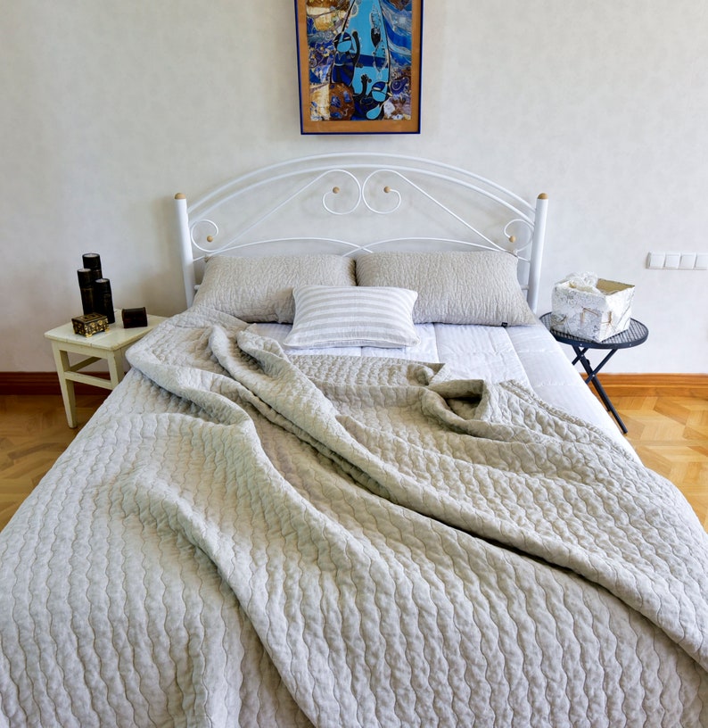K&ouml;nnte beinhalten: Ein wei&szlig;es Metallbettgestell mit einer hellgrauen Leinenbettdecke und zwei Kissen. Das Bett ist mit einem wei&szlig;en Laken und einem gestreiften Kissen bezogen. Das Bett steht in einem Zimmer mit wei&szlig;en W&auml;nden und einem Holzboden.