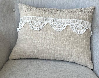 May include: A beige rectangular pillow with a quilted pattern and a white lace trim. The pillow has a decorative embroidered design along the top edge and is placed on a gray armchair.