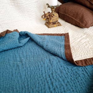 May include: A blue and brown quilted throw blanket with a white quilted blanket underneath. The throw blanket is folded over the white blanket. An ornate gold elephant figurine is on the white blanket.
