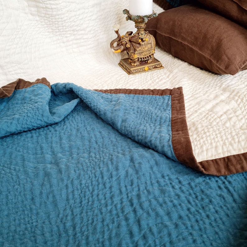 May include: A blue and brown quilted throw blanket with a white quilted blanket underneath. The throw blanket is folded over the white blanket. An ornate gold elephant figurine is on the white blanket.