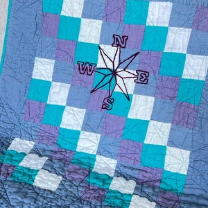 Puede incluir: Un edredón hecho a mano con un patrón de tablero de ajedrez en tonos de azul, turquesa, morado y blanco. Una rosa de los vientos está bordada en el centro, con las letras N, W, E y S. El edredón tiene una textura acolchada.