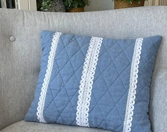 May include: A square throw pillow in a dusty blue color featuring a quilted diamond pattern across the surface. The pillow is accented with two vertical strips of white lace trim that run from the top edge to the bottom edge of the cushion.