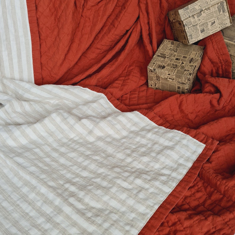 Puede incluir: Una manta doblada, a rayas, de color blanco roto y beige con un borde rojo est&aacute; superpuesta a un edred&oacute;n rojo texturizado. Dos peque&ntilde;as cajas decorativas con estampado de peri&oacute;dico son visibles en el fondo. La ropa de cama tiene un aspecto suave y acogedor.