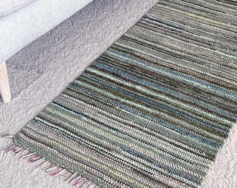 May include: A rectangular, striped rug in shades of green, blue, and brown with a fringed edge. The rug is placed on a patterned, light gray surface near a light gray sofa.