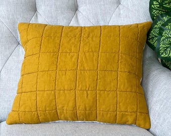May include: A rectangular, mustard-yellow pillow with a quilted grid pattern. The pillow is placed on a light gray upholstered chair. The pillow's texture and color provide a warm contrast to the neutral background.