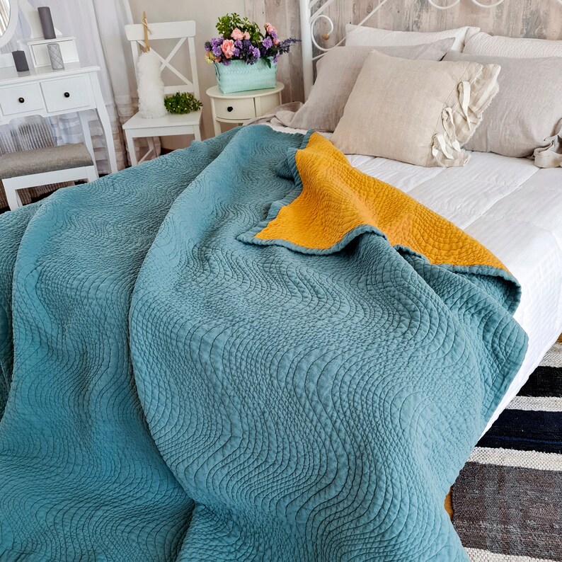 May include: A teal blue quilted throw blanket with a yellow underside. The blanket is folded on a bed with white sheets and pillows.
