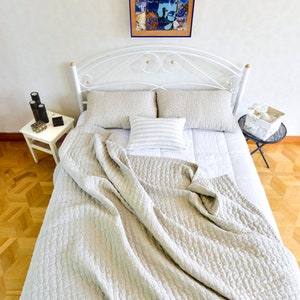 K&ouml;nnte beinhalten: Ein wei&szlig;es Metallbettgestell mit einer wei&szlig;en und grauen gesteppten Bettdecke. Das Bett ist mit zwei Kissen und einem Dekokissen mit Streifenmuster bezogen. Ein gerahmtes Kunstwerk mit blauem und orangefarbenem Design h&auml;ngt an der Wand &uuml;ber dem Bett.