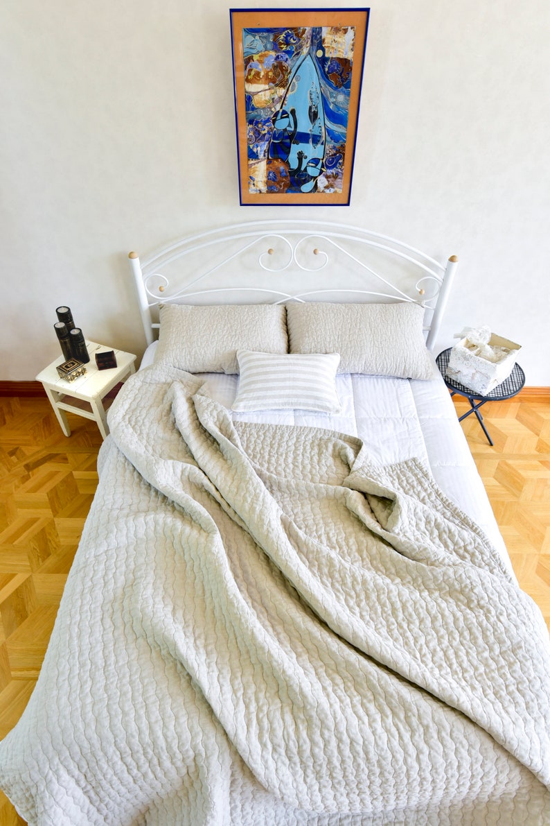 K&ouml;nnte beinhalten: Ein wei&szlig;es Metallbettgestell mit einer wei&szlig;en und grauen gesteppten Bettdecke. Das Bett ist mit zwei Kissen und einem Dekokissen mit Streifenmuster bezogen. Ein gerahmtes Kunstwerk mit blauem und orangefarbenem Design h&auml;ngt an der Wand &uuml;ber dem Bett.