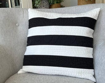 May include: A decorative pillow with a black and white striped pattern. The pillow is rectangular and has a quilted texture. The stripes are horizontal and alternate between black and white. The pillow is placed on a gray armchair.