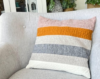 May include: A decorative pillow with horizontal stripes of pink, orange, gray, and white. The pillow is quilted and rests on a light gray armchair. The pillow adds a pop of color to the neutral-toned furniture.