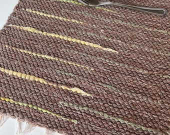 May include: A rectangular brown woven placemat featuring horizontal stripes in pale yellow and muted green. The placemat has a textured, chunky knit appearance with short white fringe along the bottom edge, topped with a white plate and a silver fork.