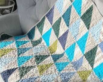 May include: A quilted throw blanket with a geometric pattern of blue, green, and gray triangles on a cream background. The blanket is draped over a gray armchair, showcasing its textured surface and detailed stitching.