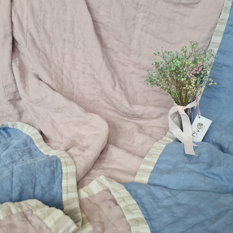Puede incluir: Una manta acolchada en tonos rosa empolvado y azul. La manta tiene un borde a rayas y un patr&oacute;n cosido. Un peque&ntilde;o ramo de flores secas en un jarr&oacute;n de cristal con una cinta rosa est&aacute; sobre la manta.