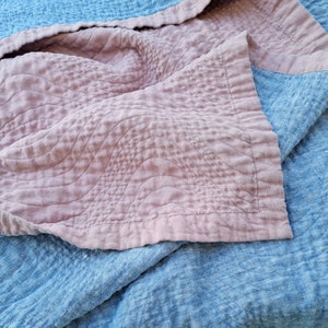 May include: A soft, pink quilted blanket with a wavy pattern, folded over a blue linen blanket.