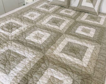 May include: A neutral-toned quilt and two matching square pillows on a bed. The quilt features a geometric pattern of squares and diamonds in shades of beige and white. The pillows have a similar design. The bed has a white metal frame.
