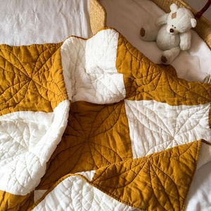 Könnte beinhalten: Eine gesteppte Decke mit einem geometrischen Muster aus abwechselnden senfgelben und weißen Quadraten. Der Stoff ist durchgehend mit einem floralen oder sternförmigen Steppmuster versehen. Ein kleiner Plüschteddy sitzt in einem Weidenkorb.