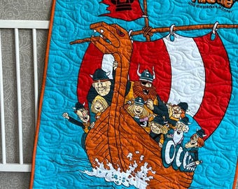 May include: A quilted baby blanket featuring a cartoon Viking ship with a crew of characters. The blanket has a blue background with an orange border and a red and white striped sail. The text "Wickie und die starken Männer" is visible.
